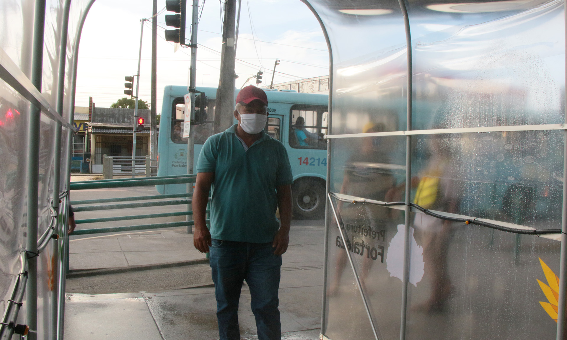 homem de boné e máscara caminhando por túnel transparente e ônibus passando ao fundo
